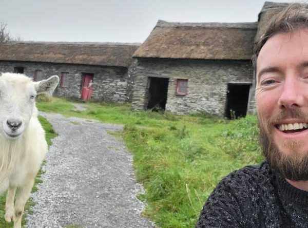 A man posing with a goat in front of a tatched cottage